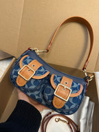 A navy blue and beige monogram shoulder bag with tan leather accents and gold-tone buckles, held by a hand inside a cardboard box.