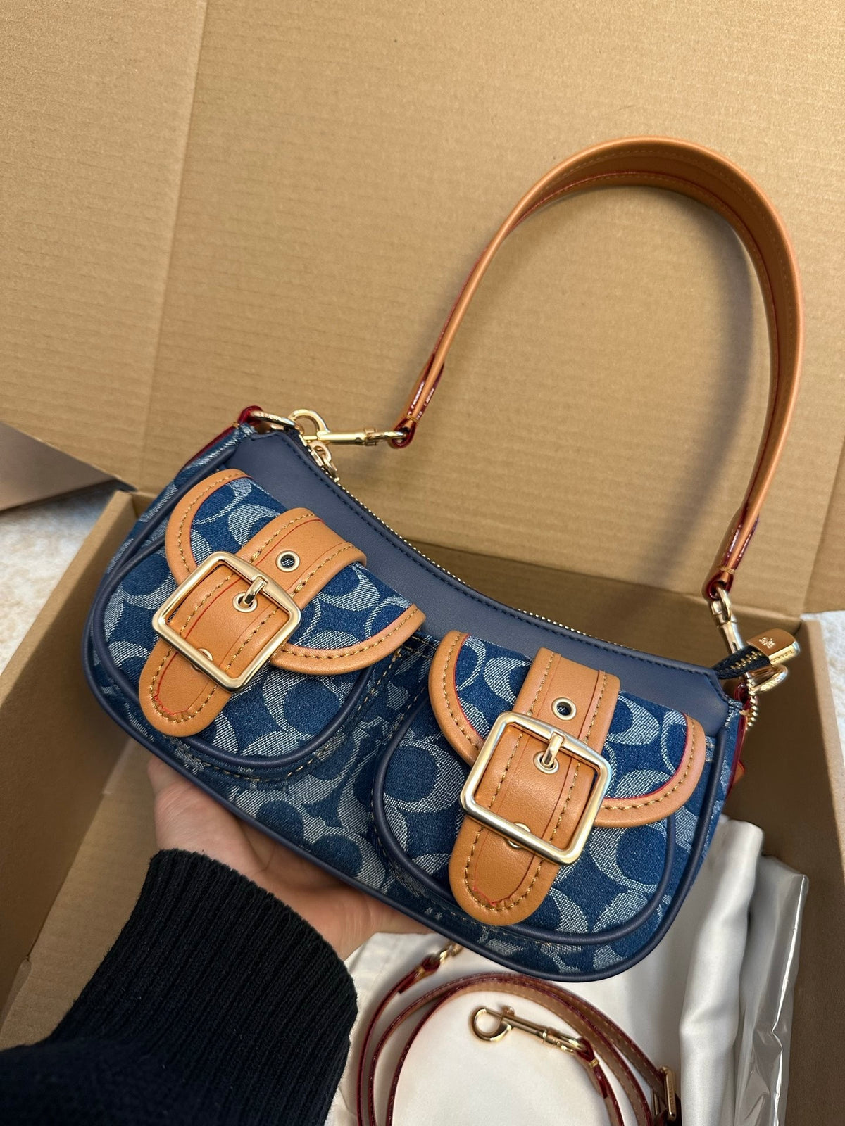 A navy blue and beige monogram shoulder bag with tan leather accents and gold-tone buckles, held by a hand inside a cardboard box.