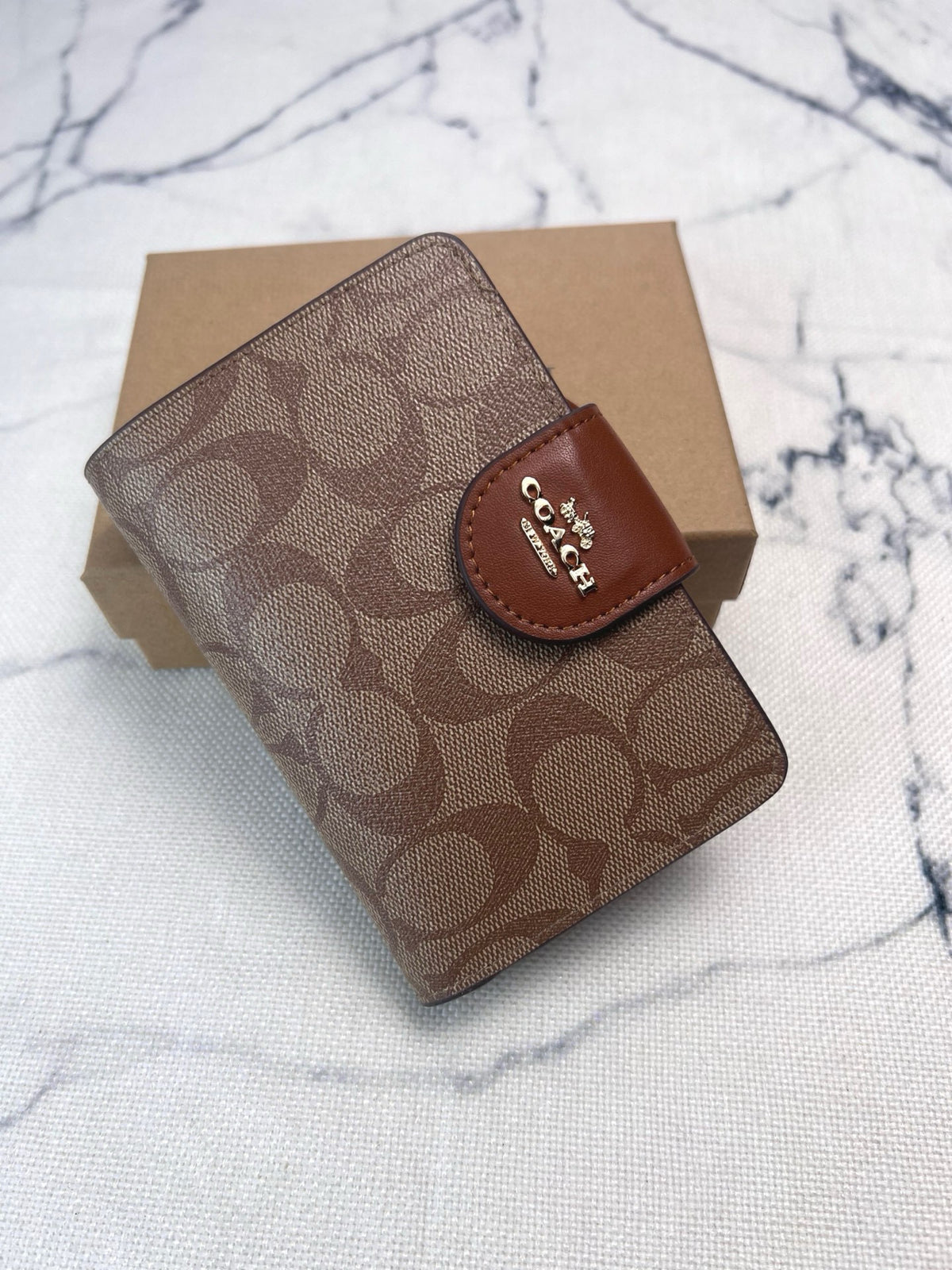 A Coach small wallet in signature brown monogram canvas with a brown leather logo tab, resting on a brown cardboard box over a white marble-patterned surface.