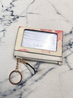 A small rectangular card holder wallet with a clear ID window, cream-colored exterior, pink floral patterned edges, and a hanging keychain, placed on a light textured fabric background.
