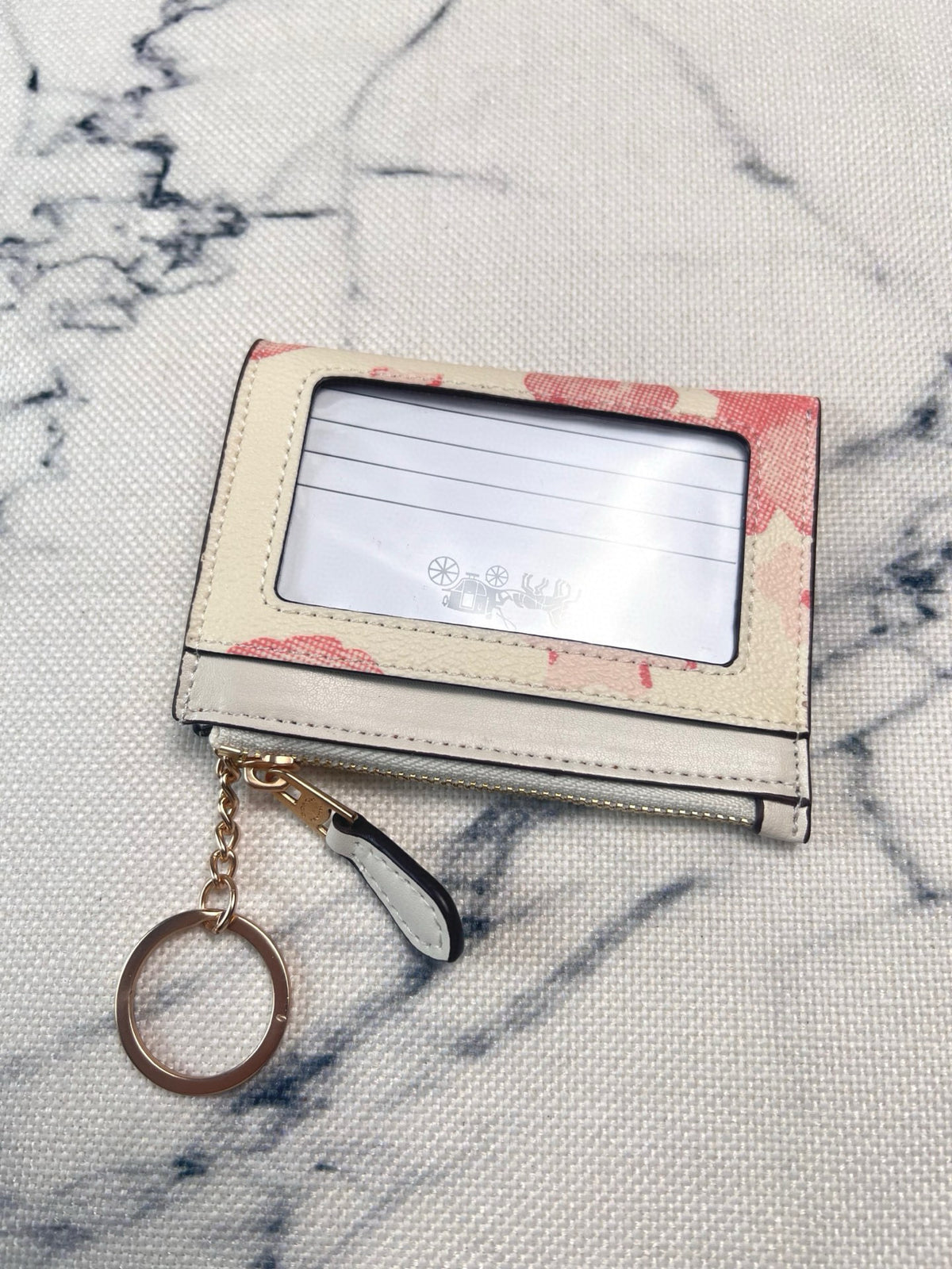 A small rectangular card holder wallet with a clear ID window, cream-colored exterior, pink floral patterned edges, and a hanging keychain, placed on a light textured fabric background.