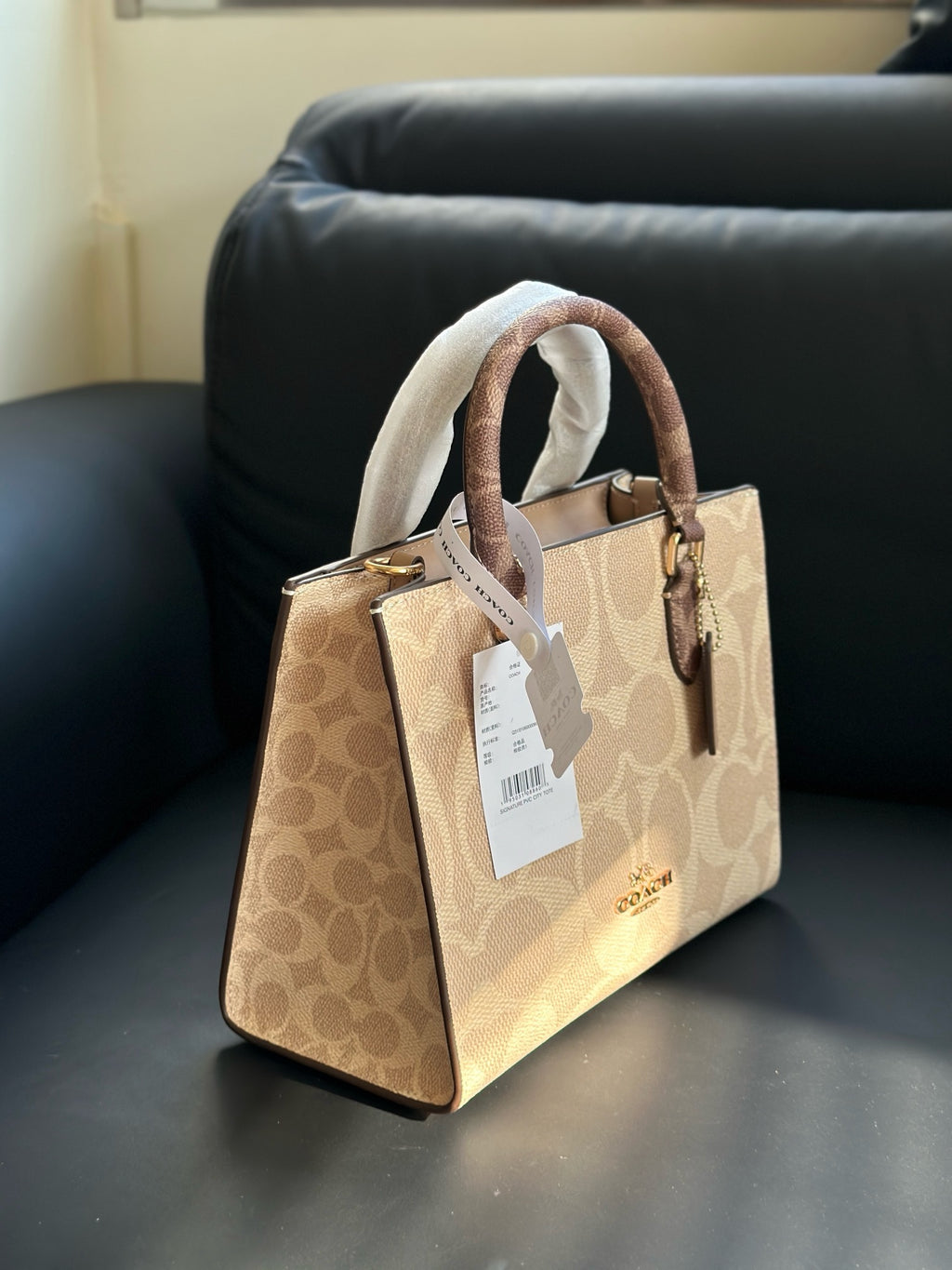 A Coach handbag in signature beige and brown monogram canvas, featuring dual top handles wrapped in white protective paper, a white hangtag, and a gold-tone logo on a black leather sofa.