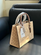 A Coach handbag in signature beige and brown monogram canvas, featuring dual top handles wrapped in white protective paper, a white hangtag, and a gold-tone logo on a black leather sofa.