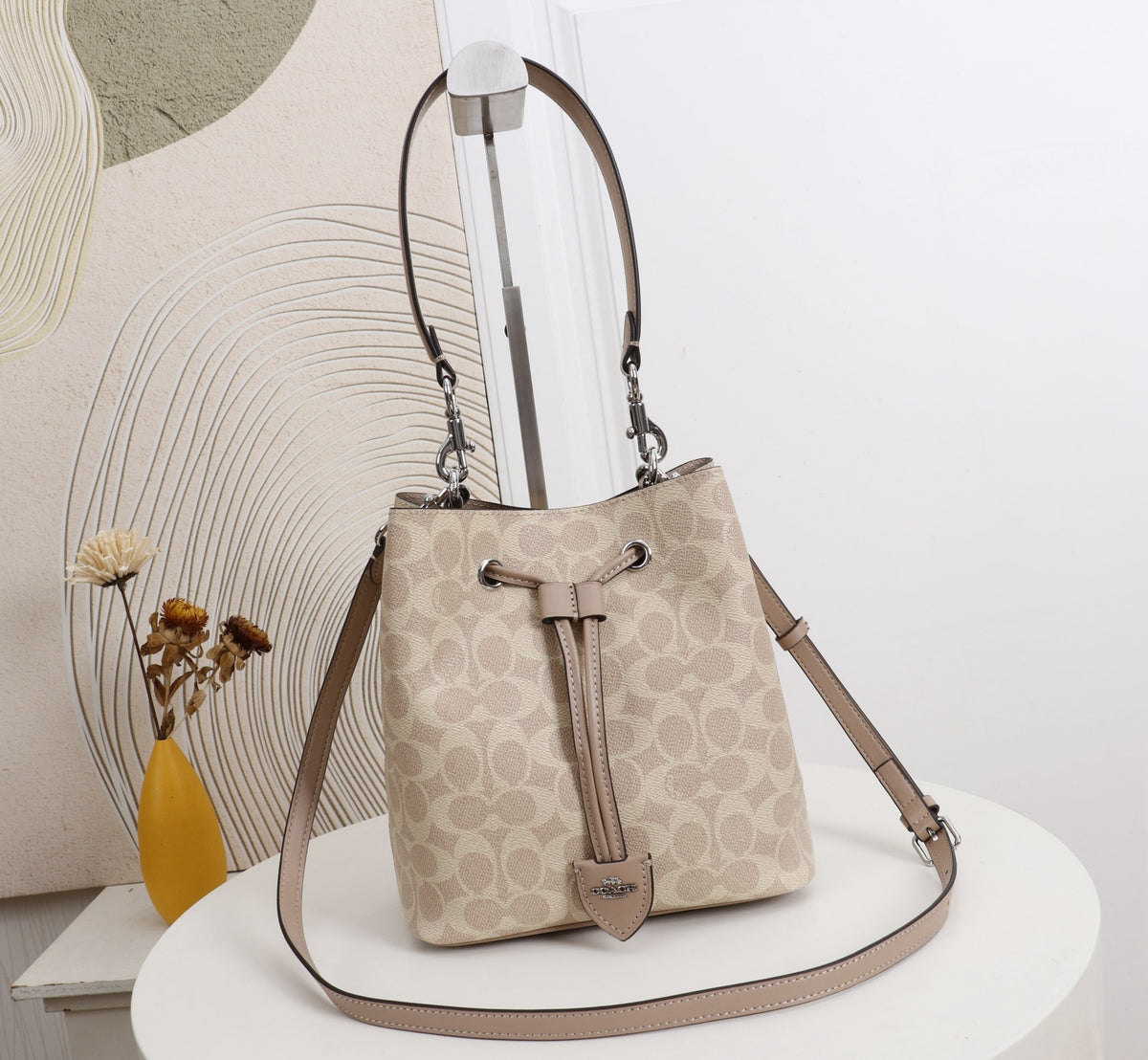 A beige and taupe monogram bucket bag with a leather strap and drawstring closure, displayed on a white round table against a modern wall with abstract line art.