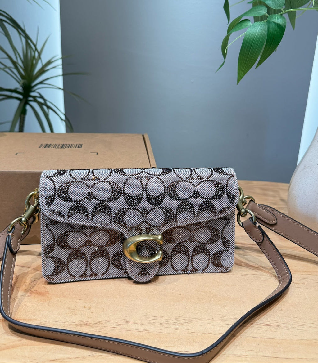 A Coach flap shoulder bag in the signature monogram canvas with a gold-tone C logo clasp, resting on a light wooden table with its box and strap visible.