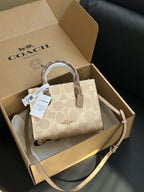 A Coach handbag in beige signature canvas sits inside an open Coach box on a dark leather surface. The box lid shows the Coach logo, and the bag has a top handle, detachable shoulder strap, and hangtag.
