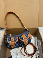 A blue and tan monogram shoulder bag with gold-tone hardware, displayed inside an open cardboard box with its dust bag and accessories.