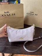 A Coach crescent-shaped shoulder bag in pale pink quilted leather with gold-tone hardware, held by a hand in front of two Coach shopping bags on a dark wooden table.