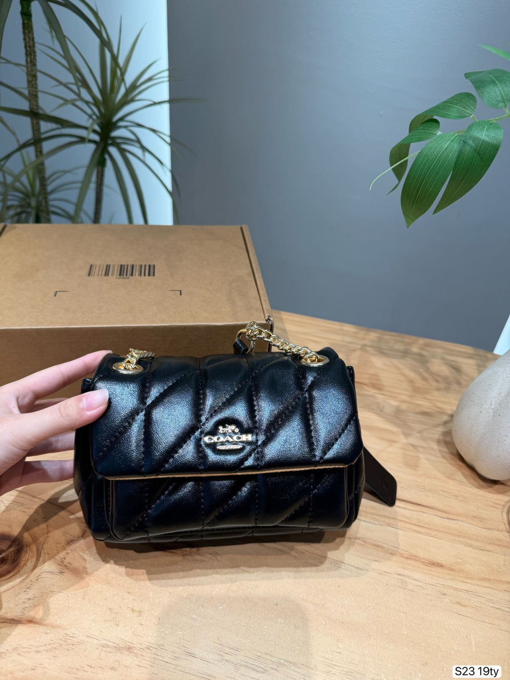 A black quilted Coach shoulder bag with gold hardware and a gold-tone logo plaque on the front, held by a hand in front of its brown cardboard box on a wooden table.