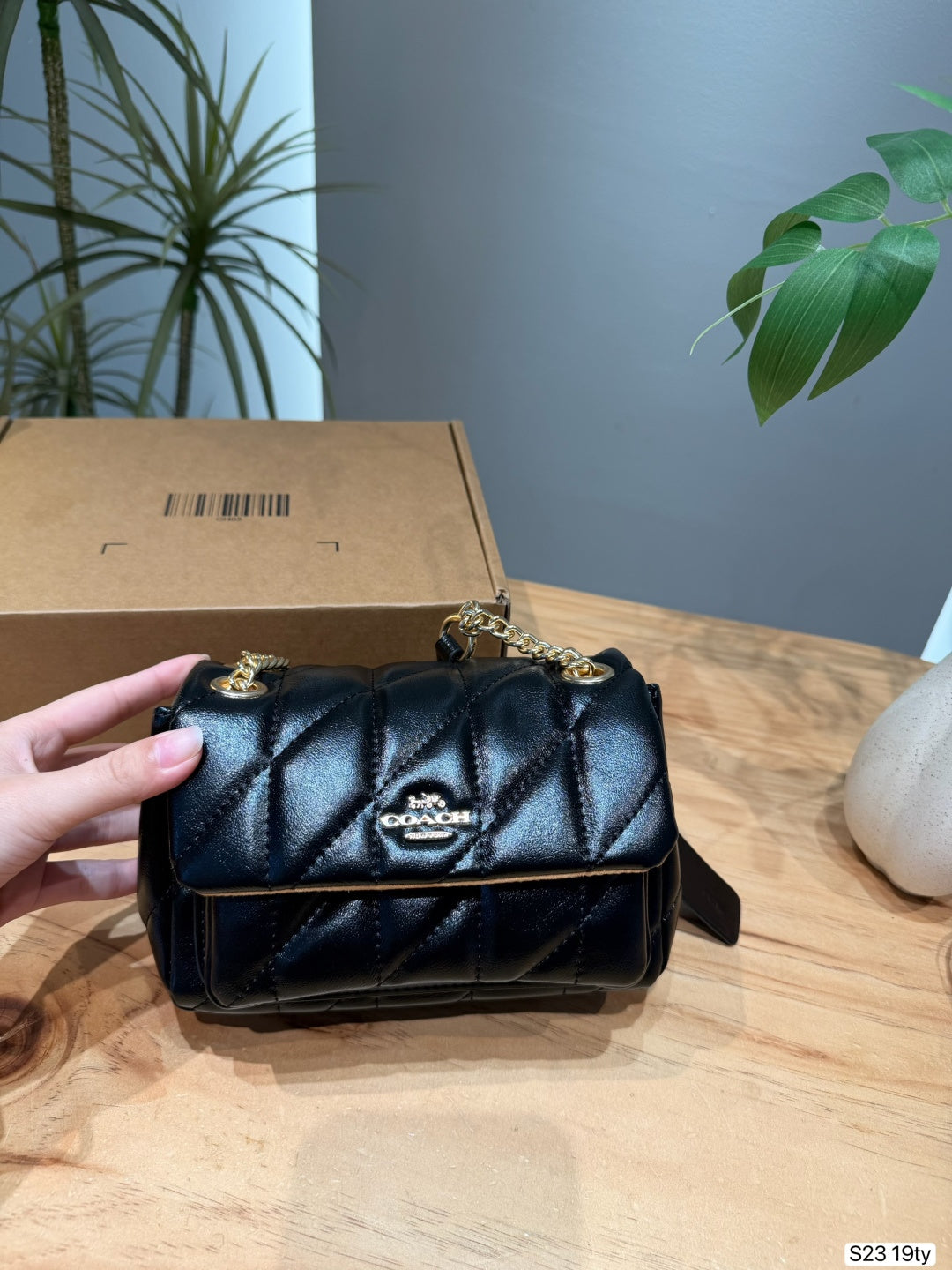 A black quilted Coach shoulder bag with gold hardware and a gold-tone logo plaque on the front, held by a hand in front of its brown cardboard box on a wooden table.
