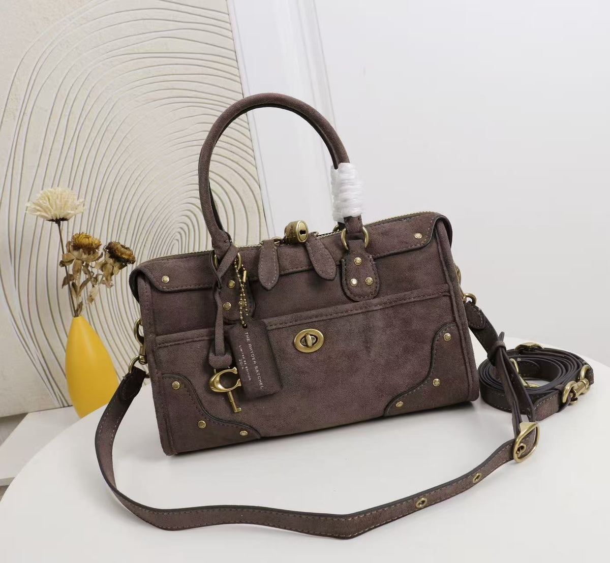 A dark brown suede handbag with gold-tone hardware, a top handle, and a detachable shoulder strap, displayed on a white surface with a decorative background.
