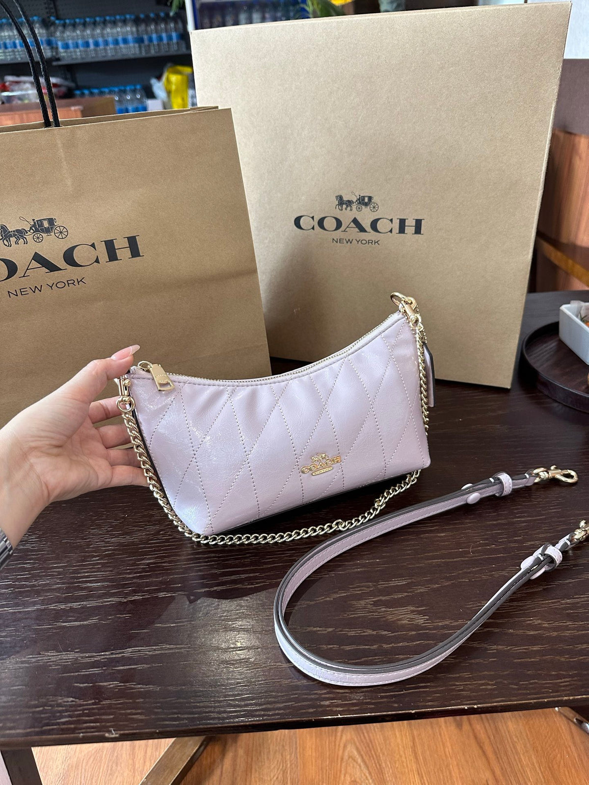 A Coach handbag in pale pink quilted leather with gold-tone hardware, held by a hand in front of a Coach shopping bag and box on a dark wooden table.