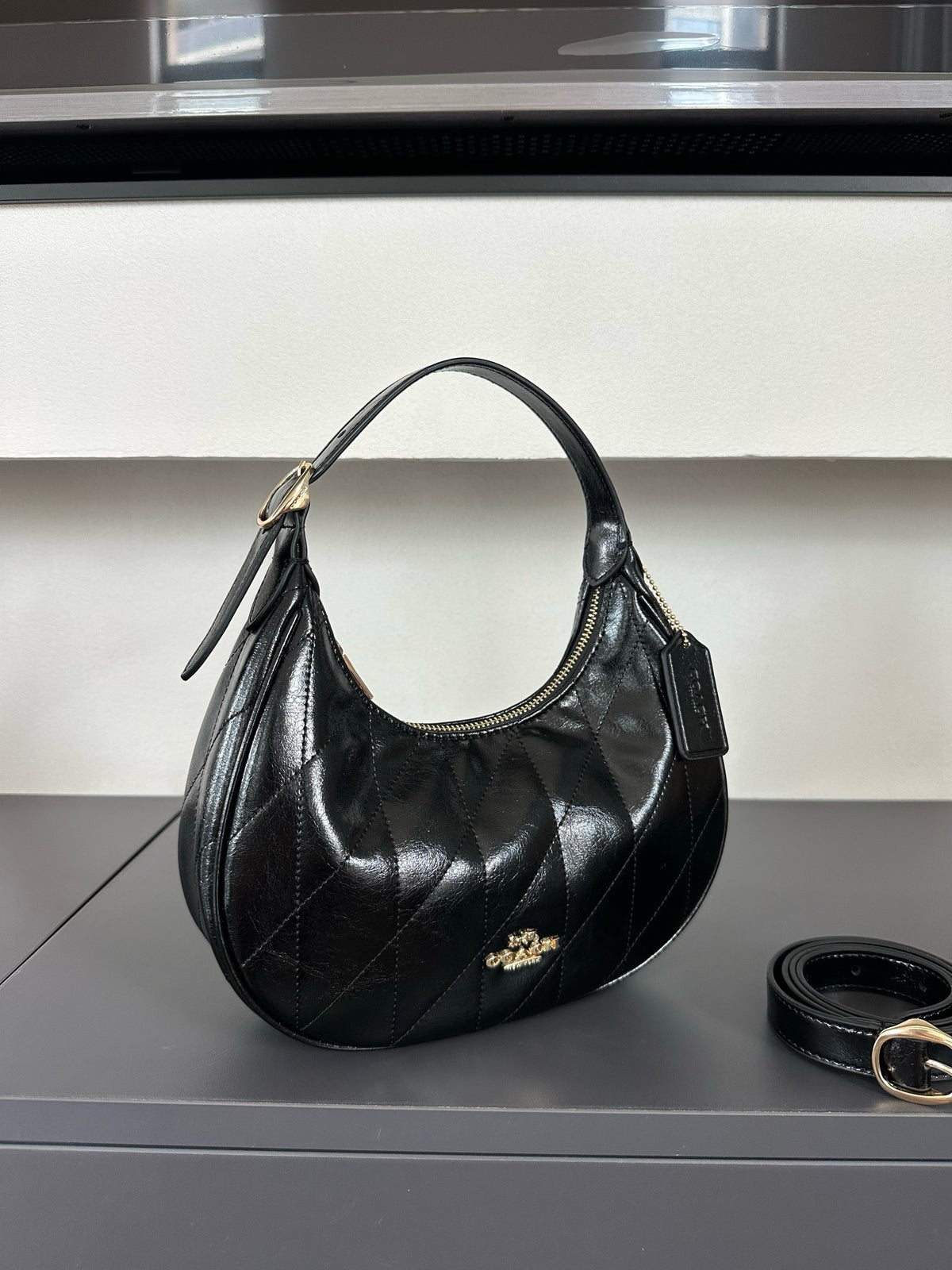 A black quilted leather shoulder bag with gold-tone hardware and a small gold logo emblem, displayed on a dark gray surface with a matching black belt nearby.