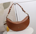 A brown crescent-shaped shoulder bag with gold-tone hardware and a small gold logo on the front, displayed on a white pedestal in front of a white wall and abstract artwork.