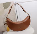 A brown crescent-shaped shoulder bag with gold-tone hardware and a small gold logo on the front, displayed on a white pedestal in front of a white wall and abstract artwork.