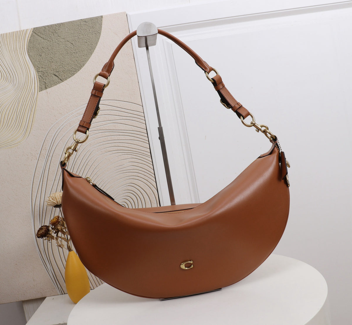 A brown crescent-shaped shoulder bag with gold-tone hardware and a small gold logo on the front, displayed on a white pedestal in front of a white wall and abstract artwork.