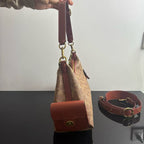 A beige coated canvas handbag with brown leather trim and gold-tone hardware, held by a hand over a dark reflective table. A matching brown leather shoulder strap with a gold buckle lies on the table to the right.