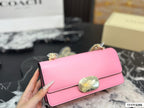 A pink Coach flap bag with a gold-tone floral clasp and chain strap, held by a hand over a black and white marble surface with Coach packaging in the background.