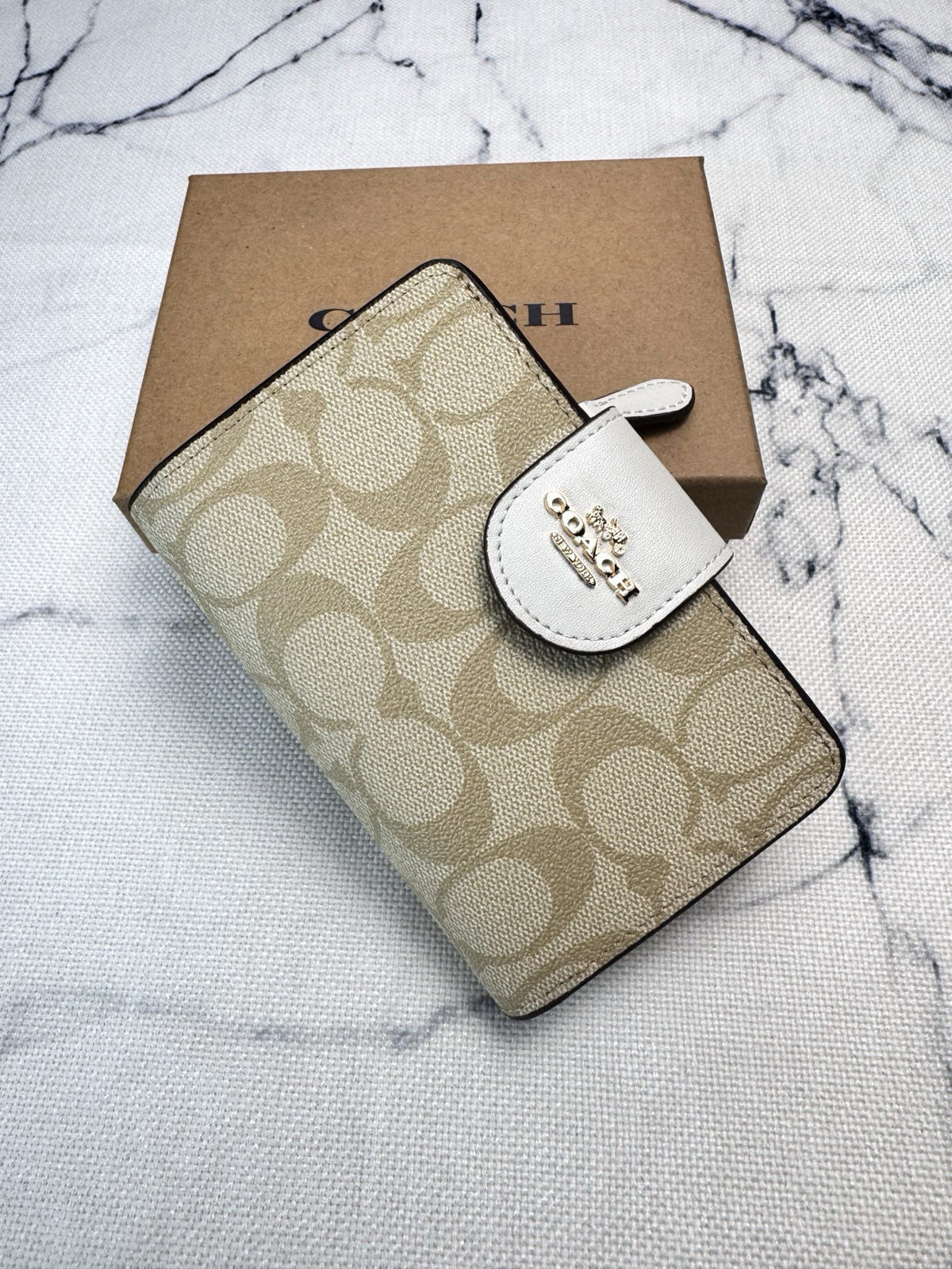 A Coach wallet in the brand's signature beige and tan monogram canvas, featuring a white leather accent tab with the Coach logo, displayed on top of its brown cardboard box against a white marble background.