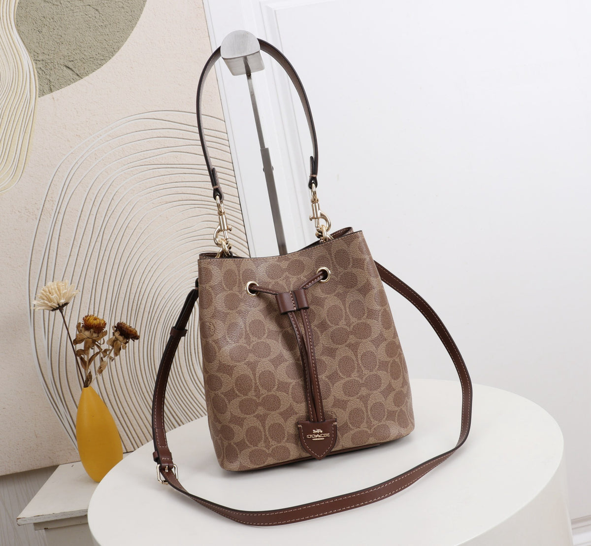 A brown and beige Coach monogram bucket bag with a drawstring closure and a long shoulder strap, displayed on a white round table in a styled interior setting.