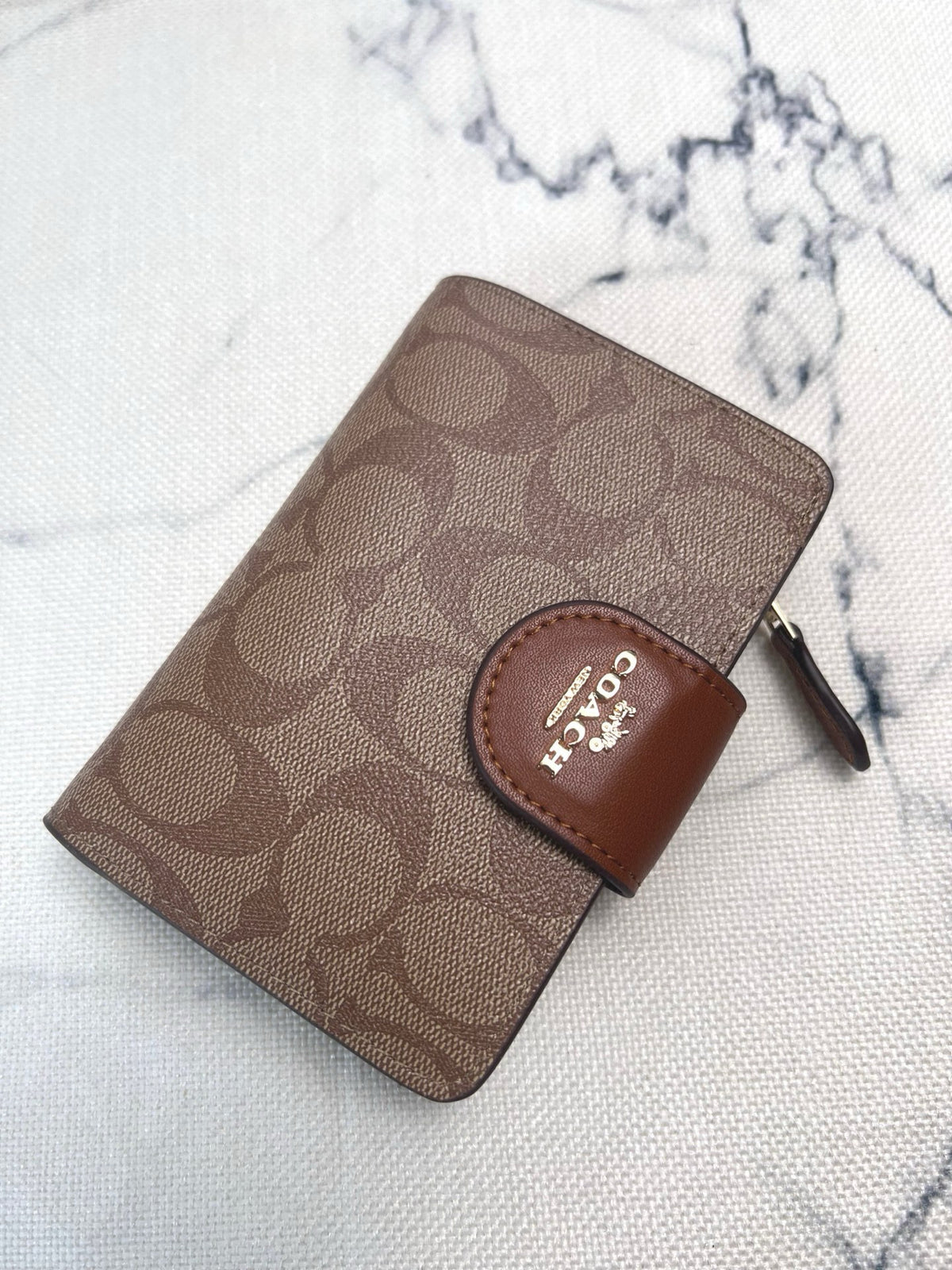 A Coach zip-around wallet in brown signature canvas with a brown leather logo tab, photographed on a light marble-patterned surface.