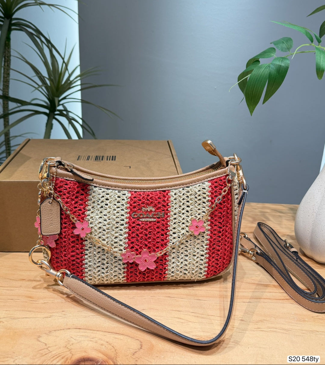 A crocheted shoulder bag with red and cream vertical stripes, decorated with pink flower appliqués and a gold-tone chain strap, displayed on a wooden table next to its brown cardboard box.
