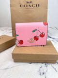 A pink Coach wallet with a cherry print, displayed on top of a brown cardboard box with a Coach bag and box in the background.