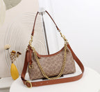A Coach hobo shoulder bag in signature monogram canvas with brown leather trim and gold-tone chain and strap hardware, displayed on a white pedestal in a styled interior setting.