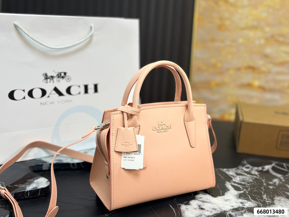 A peach-pink Coach handbag with a top handle, leather strap, and embossed logo, held by a hand with silver nail art. A white Cozzi tag is attached, and a cardboard box is visible in the blurred background.