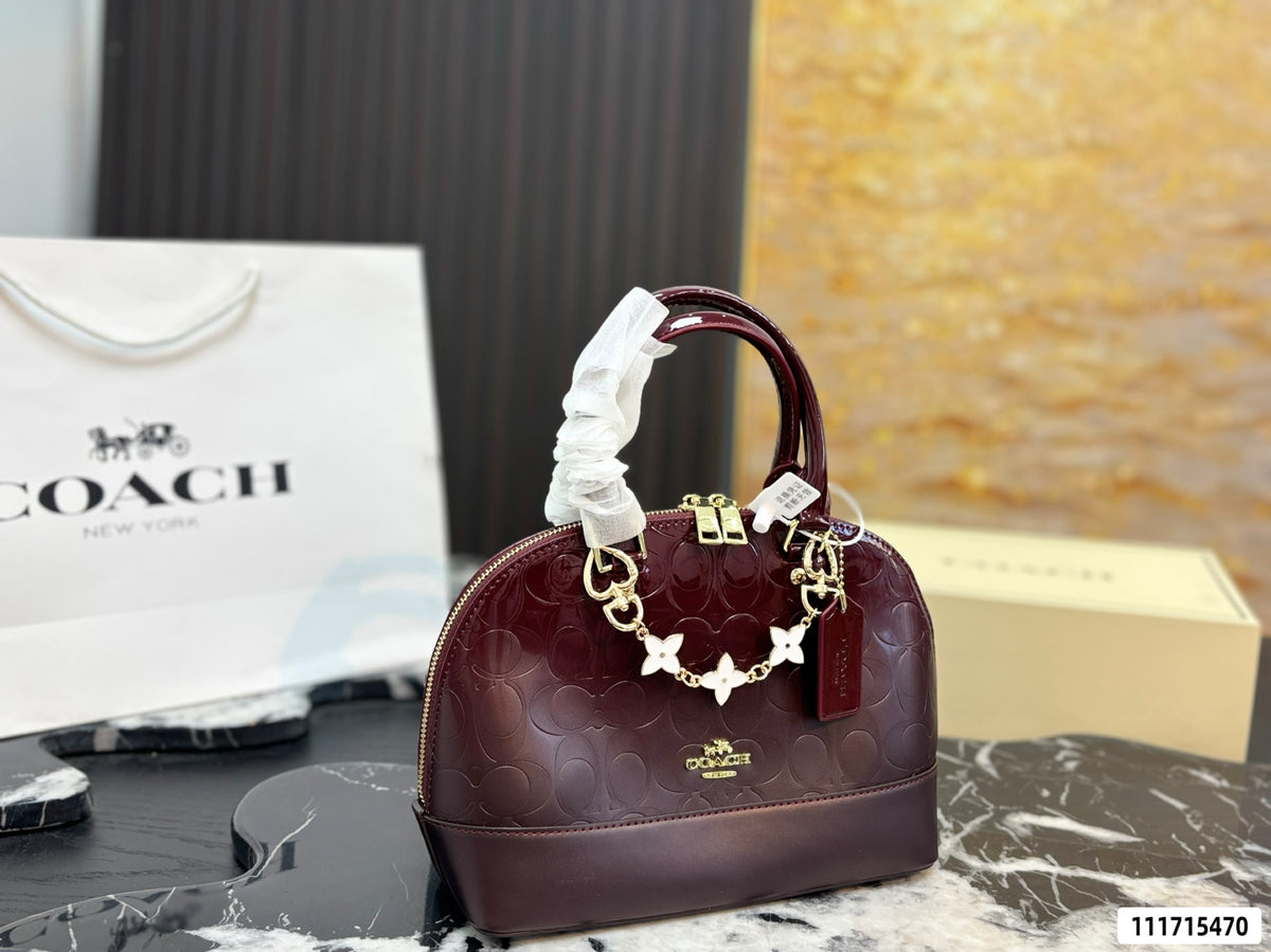 A Coach handbag in deep burgundy with the signature embossed logo pattern, gold-tone hardware, and a white protective paper sleeve on the handle, displayed on a marble surface with a Coach shopping bag and box in the background.