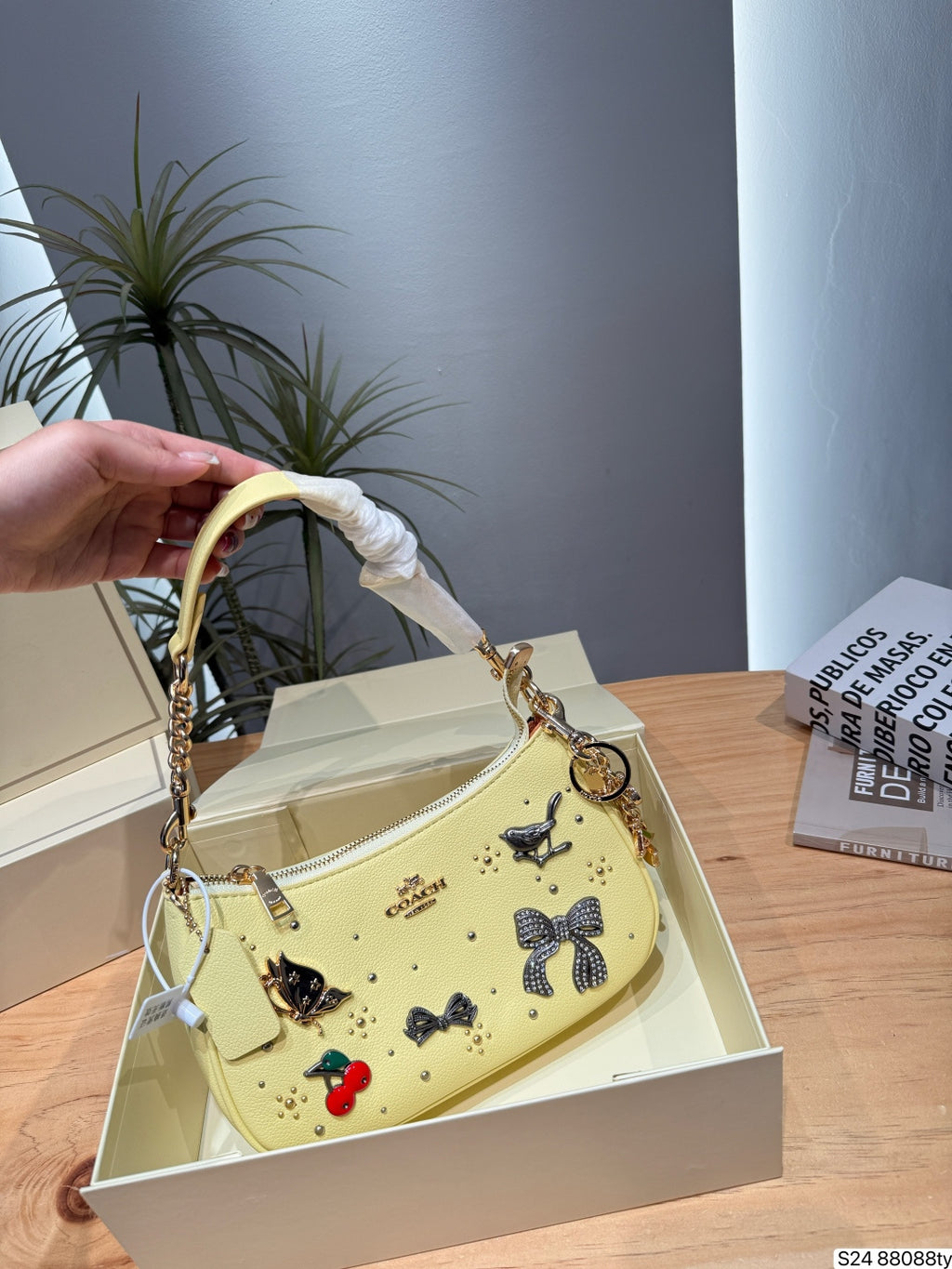 A pale yellow Coach handbag with decorative bows, cherries, and gold hardware, held by a hand over an open cream-colored box on a wooden table.
