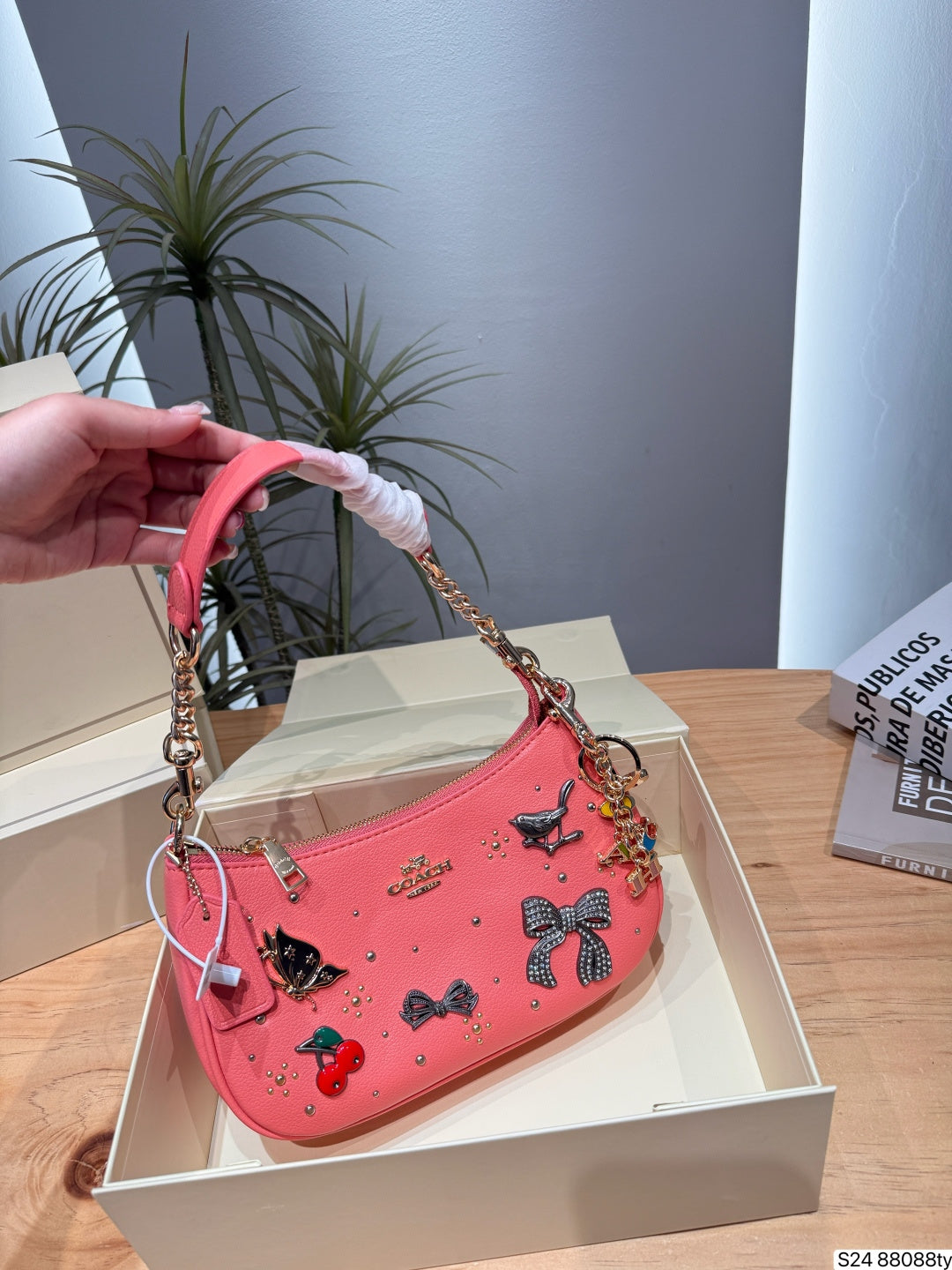 A pink shoulder bag with decorative bows, cherries, and rhinestones, held by a hand over an open beige box on a wooden table.