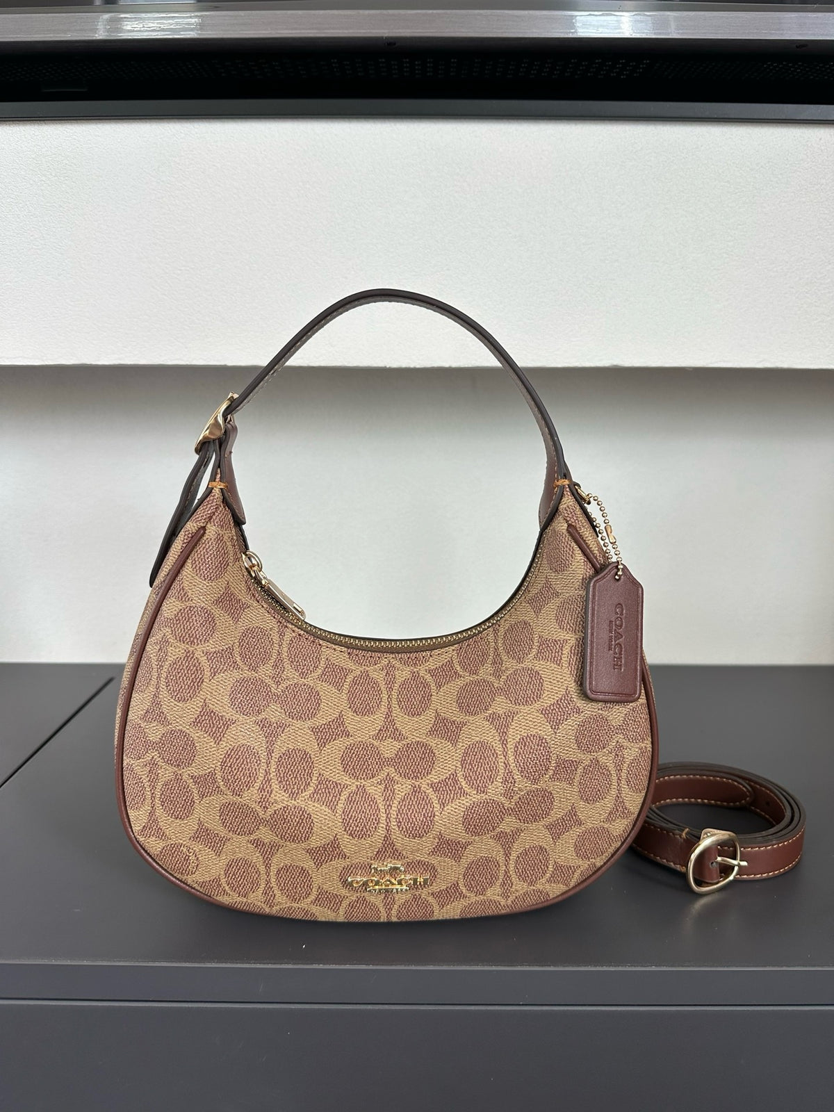 A Coach hobo shoulder bag in the classic monogram canvas, featuring a brown leather handle and a matching detachable shoulder strap, displayed on a dark gray surface against a white paneled background.