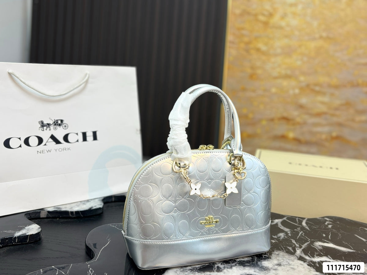 A Coach New York metallic silver dome handbag with the signature monogram pattern, gold-tone hardware, and a white fabric wristlet wrap on the handle. A white Coach shopping bag and a beige Coach box are visible in the background on a dark marble surface.