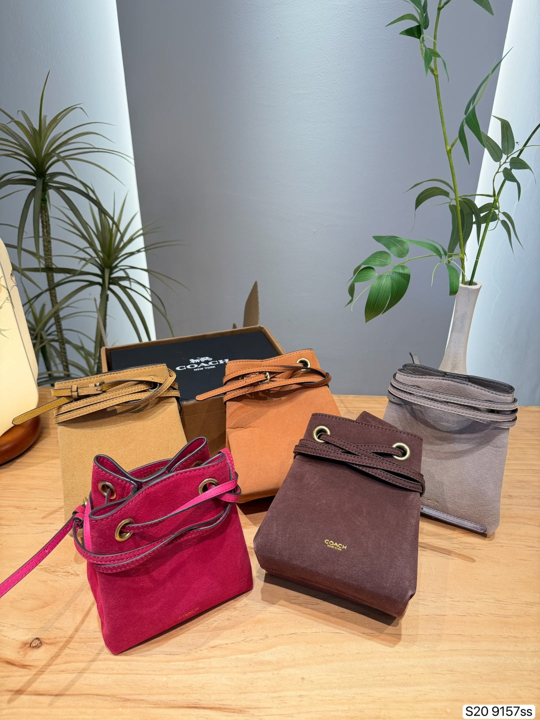 Four Coach drawstring pouches in pink, brown, tan, and gray, displayed on a wooden table with their branded boxes and a Coach box in the background, accented by indoor plants.