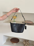 A black leather shoulder bag with gold-tone hardware and a small gold logo on the front, held by a person's hand in front of a white shelf with decorative items.
