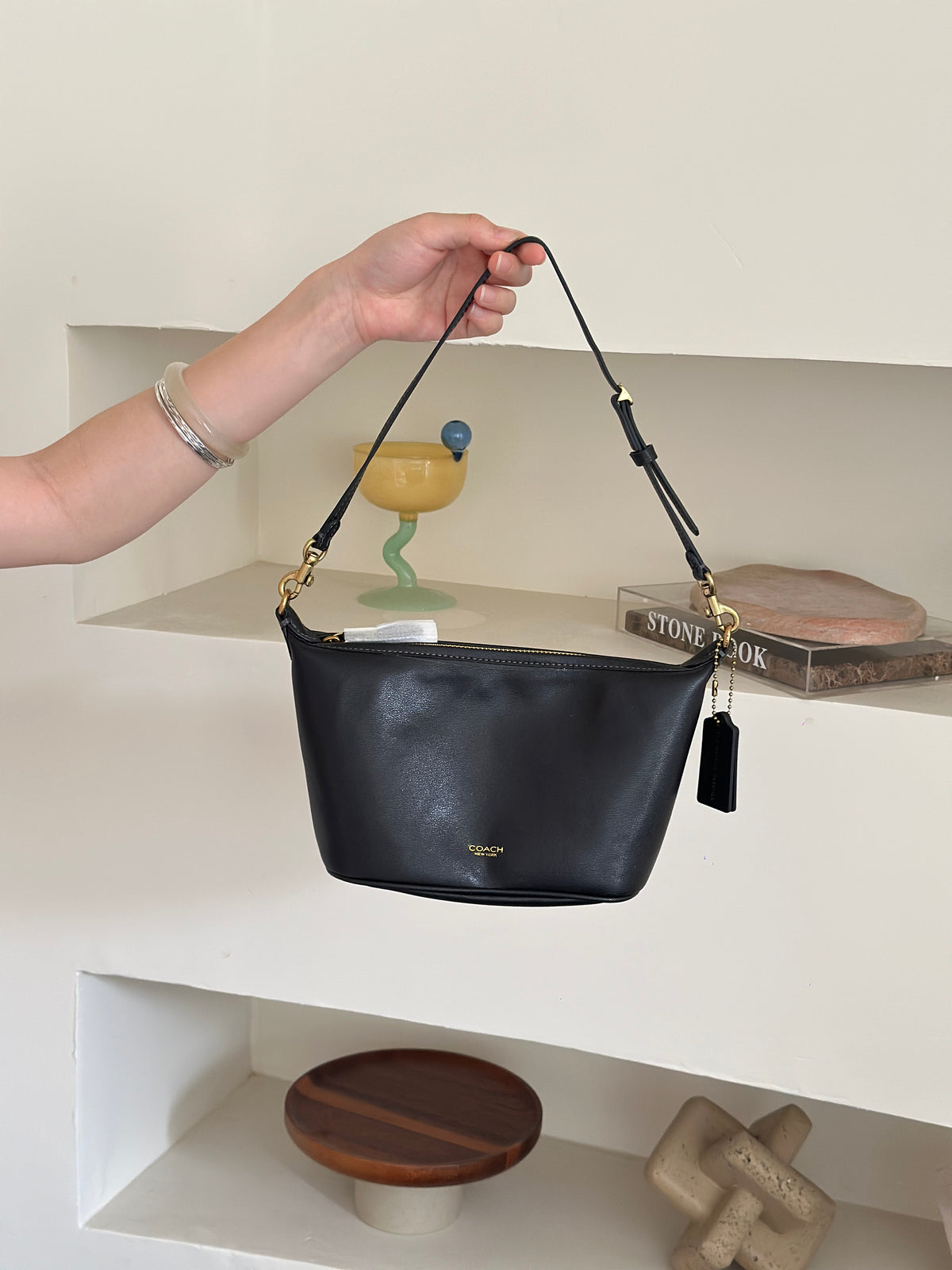 A black leather shoulder bag with gold-tone hardware and a small gold logo on the front, held by a person's hand in front of a white shelf with decorative items.