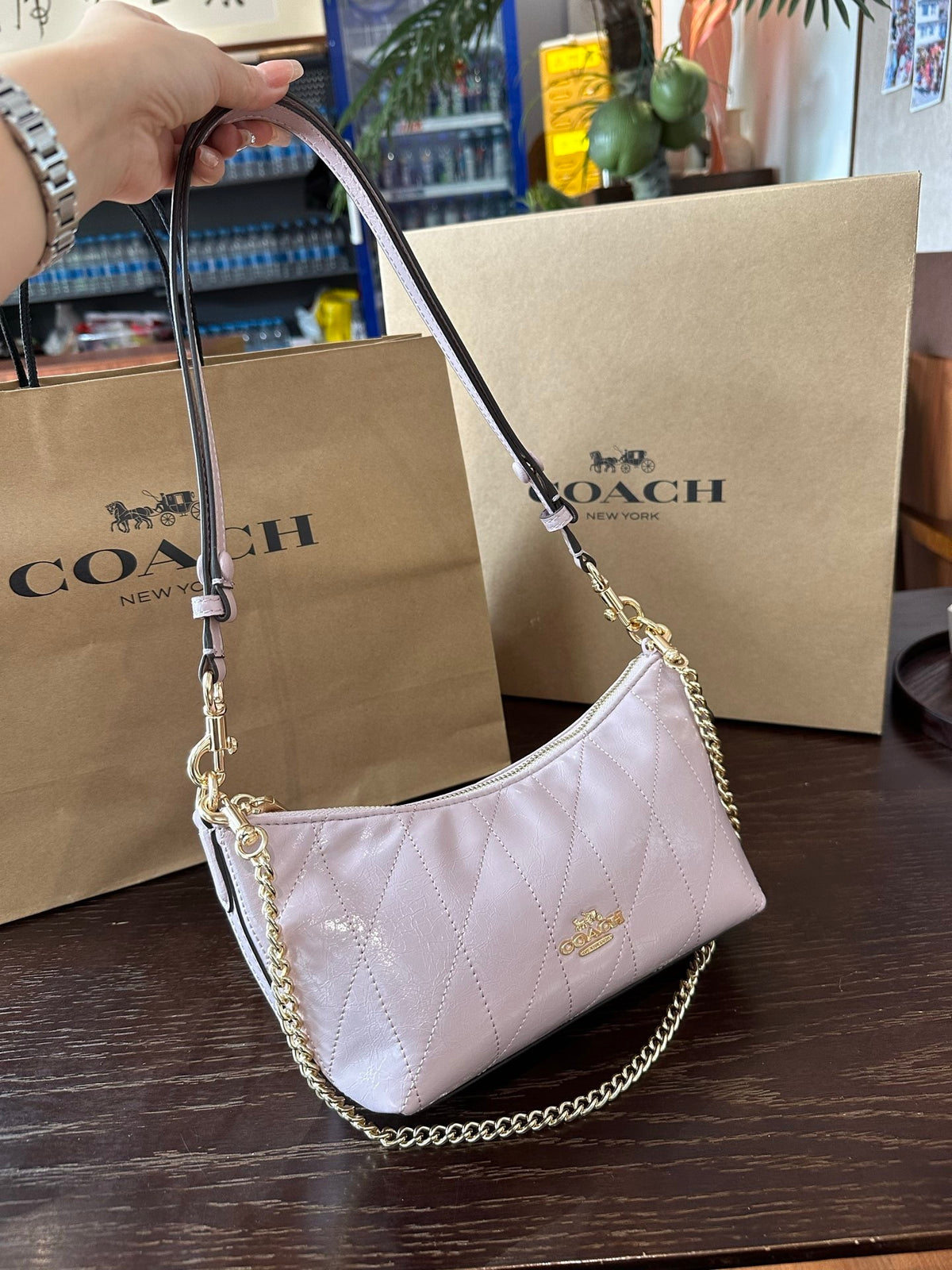 A Coach shoulder bag in pale pink quilted leather with gold-tone hardware and chain strap, held by a hand in front of Coach shopping bags on a wooden table.