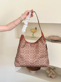 A person's arm holds up a Coach hobo bag with a brown leather shoulder strap and a white sheer scrunchie attached to the strap. The bag features the signature Coach monogram pattern in brown and beige.