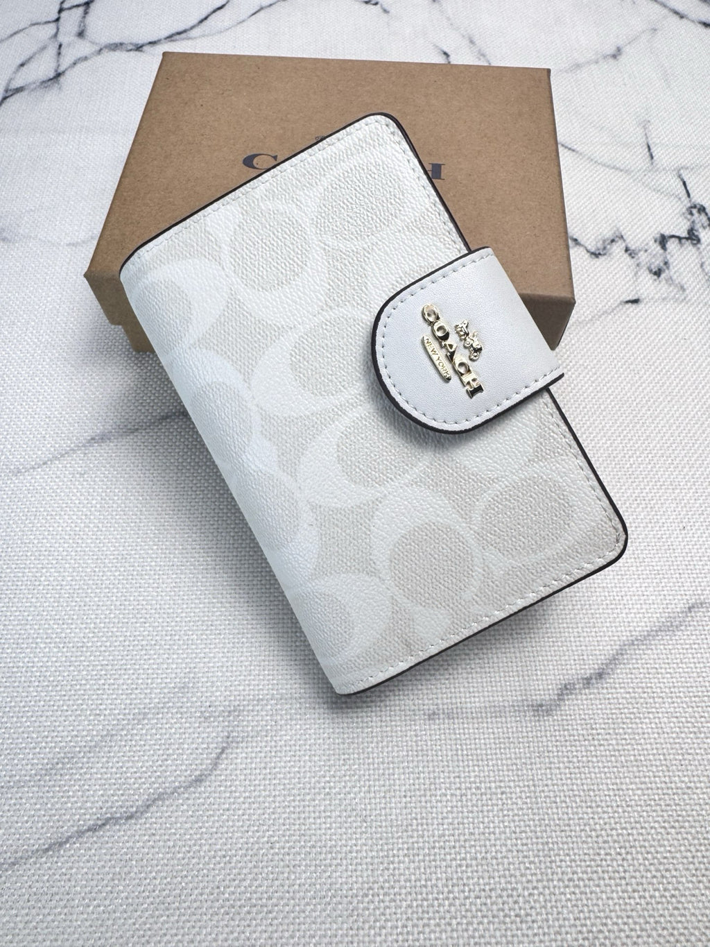 A Coach small wallet in light gray signature monogram canvas with a white leather magnetic tab closure featuring a gold-tone Coach logo, shown with its brown cardboard box on a light marble-textured surface.