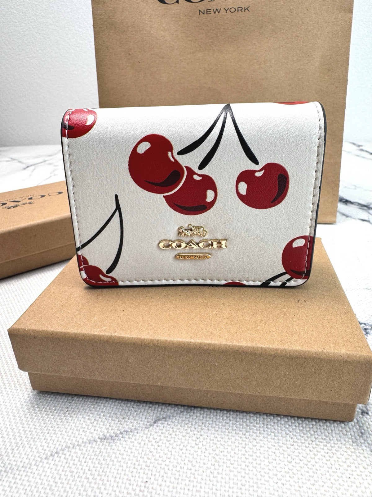 A Coach wallet with a white faux leather body decorated with red cherries and black stems, resting on a brown cardboard box with the Coach logo visible on the box and a shopping bag in the background.