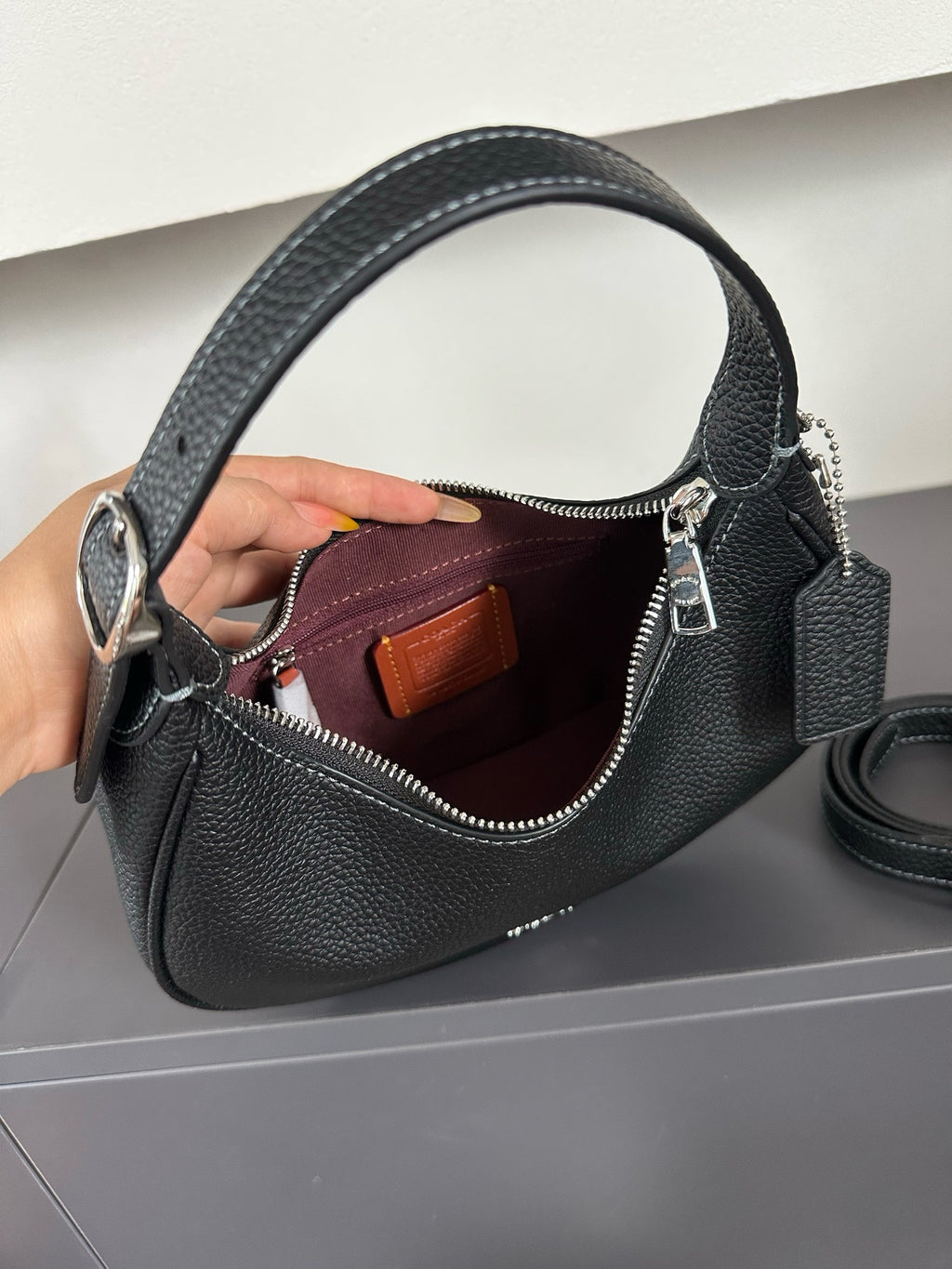 A black pebbled leather handbag with a top handle, shown open with a hand holding it to display the interior. The bag features silver-tone hardware, a zippered top, a small brown leather interior label, and a matching black leather tag with a silver clasp.